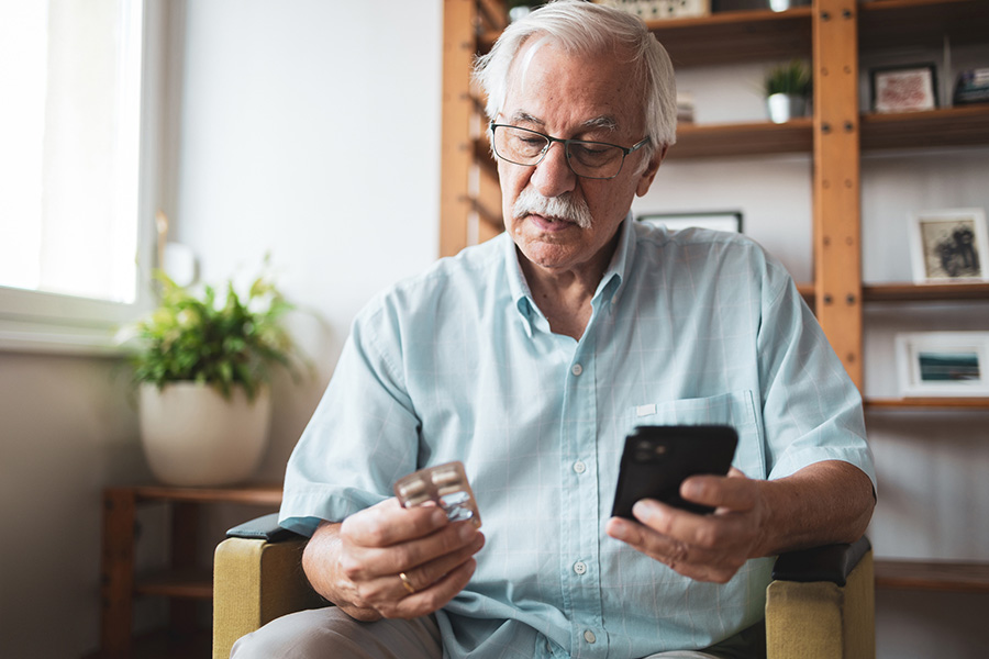 Senior adult man sitting in a chair, holding a blister pack and using a smartphone, possibly checking medication instructions or health information online