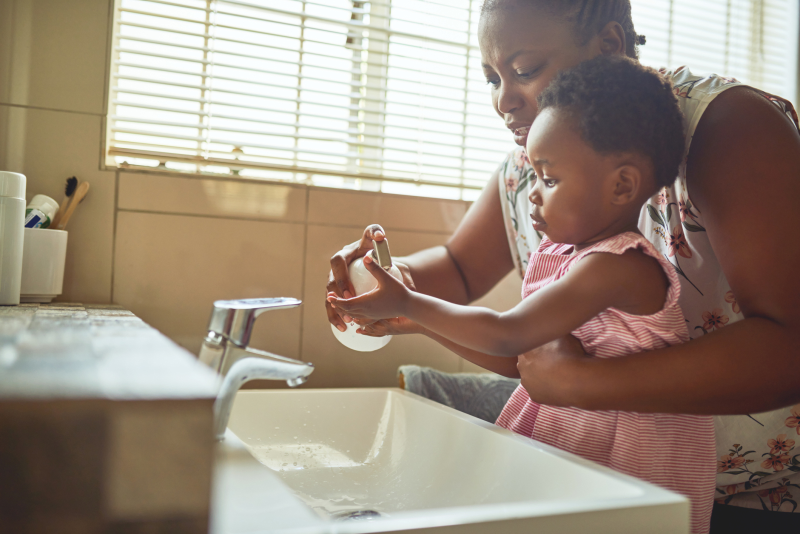 A mother holding a soap dispenser teachers her young daughter how to wash her hands at the bathroom sink
