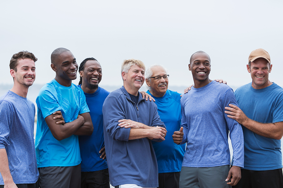 A diverse group of six men of different ages and backgrounds stand together outdoors, smiling and supporting one another with arms around shoulders, representing friendship, community, and men's health awareness.