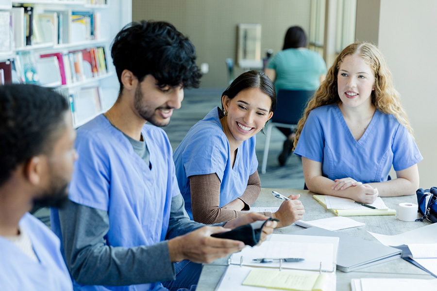 Nursing students sit at a table with papers and books strewn about, smiling and working on a class project together