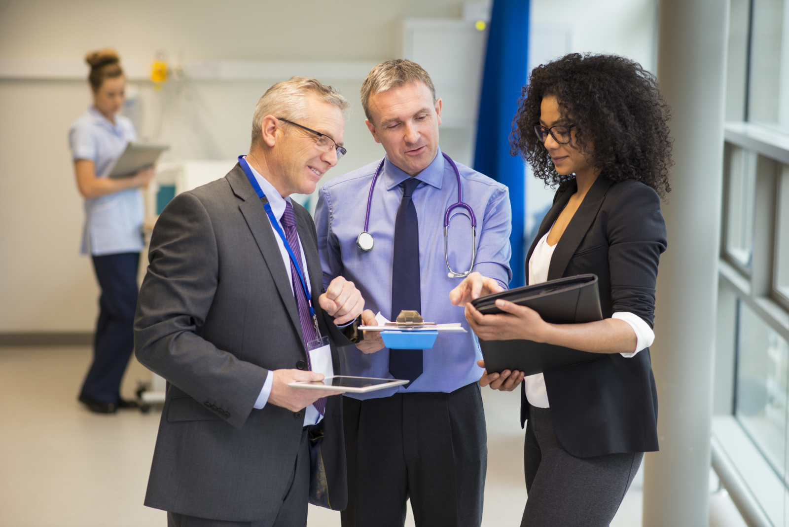 Medical administrators in suits conversing un a clinic while holding clipboards and portfolios. Medical staff walk by in the background.