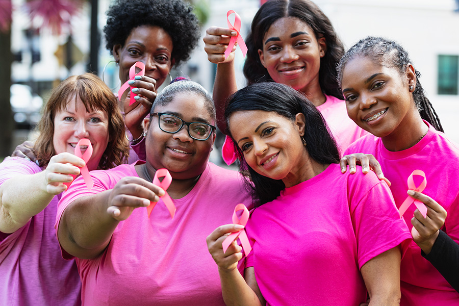 A woman stands outdoors, smiling with a pink ribbon pinned to her shirt, showing support for breast cancer awareness.